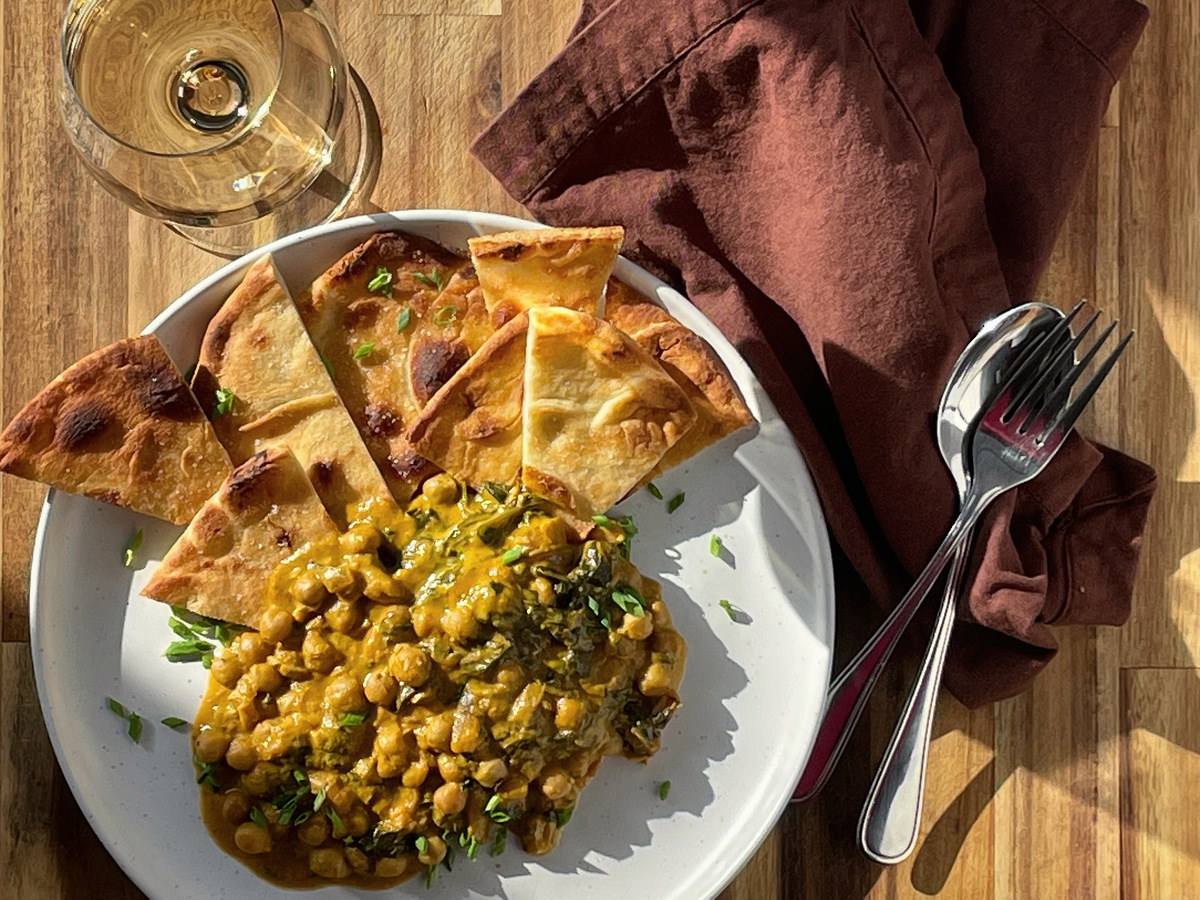A white plate with pumpkin and chickpea curry and toasted naan sitting on a wood board beside a glass of white wine and brown linen napkin